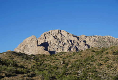 A rugged mountain peak rises against a clear blue sky, surrounded by green shrubs and rocky terrain.