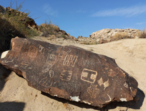 Petroglyphs etched on a large rock in a desert landscape, surrounded by sandy terrain and sparse vegetation.