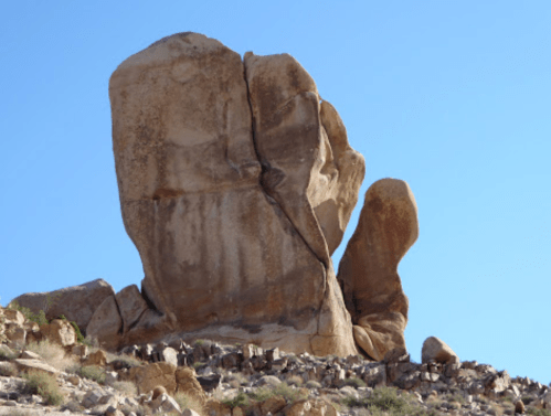 A large, uniquely shaped rock formation stands against a clear blue sky, surrounded by smaller rocks and desert landscape.