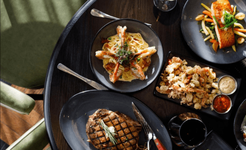 A table set with a steak, shrimp pasta, fried seafood, and a colorful vegetable dish, with drinks in the background.