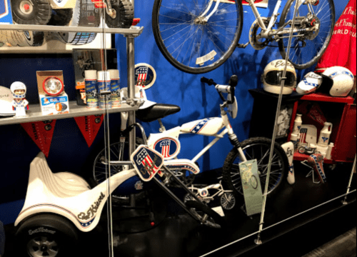 Display of vintage bicycles, helmets, and memorabilia in a glass case, featuring a white bike with red and blue accents.