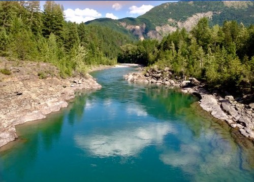 A serene river flows through a lush green landscape, surrounded by rocky banks and mountains under a blue sky.