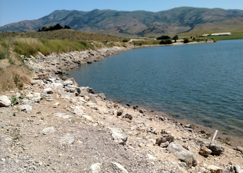 A rocky shoreline beside a calm lake, with mountains in the background under a clear blue sky.