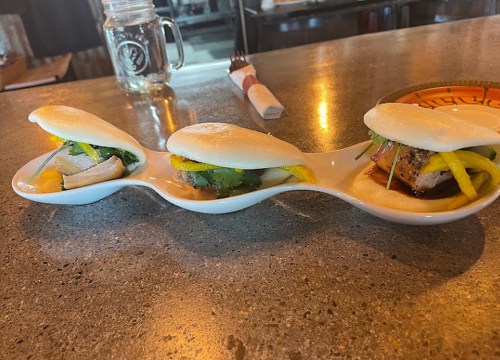 Three bao buns filled with various ingredients, served on a curved white plate, on a restaurant countertop.