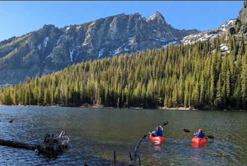 Two kayakers in red kayaks paddle on a calm lake surrounded by tall pine trees and mountains in the background.