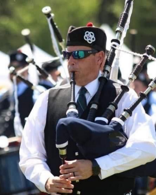 A man in sunglasses plays the bagpipes, wearing a black vest and hat, with other musicians in the background.