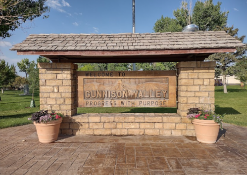 Welcome sign for Gunnison Valley, featuring stone structure and potted plants, set in a green park area.