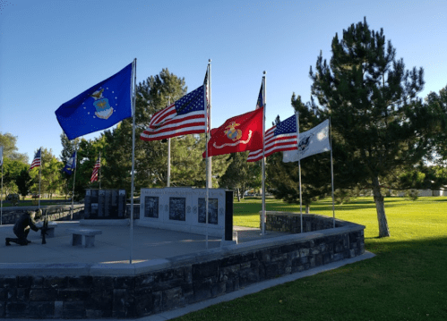 A memorial with multiple flags, including U.S. and military flags, surrounded by trees in a park setting.