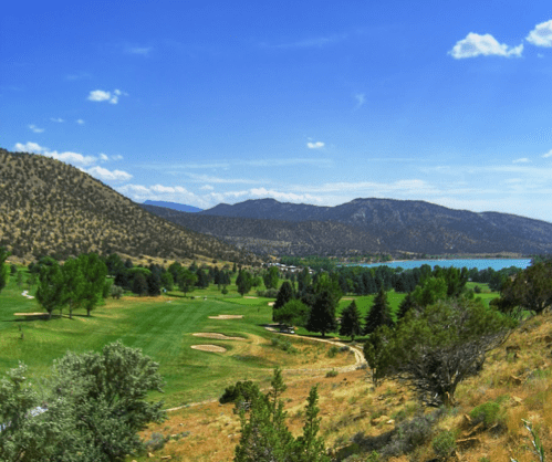 Scenic view of a golf course with rolling hills, trees, and a lake under a clear blue sky.