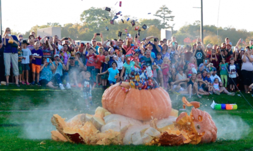 A large pumpkin explodes, showering candy over a cheering crowd at a festive outdoor event.
