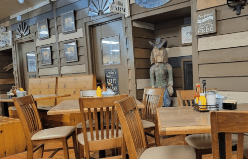 A rustic restaurant interior with wooden tables, chairs, and a cowboy statue against a wooden wall.