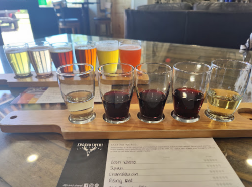 A wooden flight board with six glasses of wine and beer samples, alongside a tasting notes sheet.