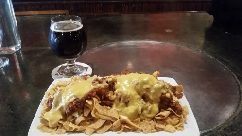 A plate of nachos topped with chili and melted cheese, alongside a glass of dark beer on a bar counter.