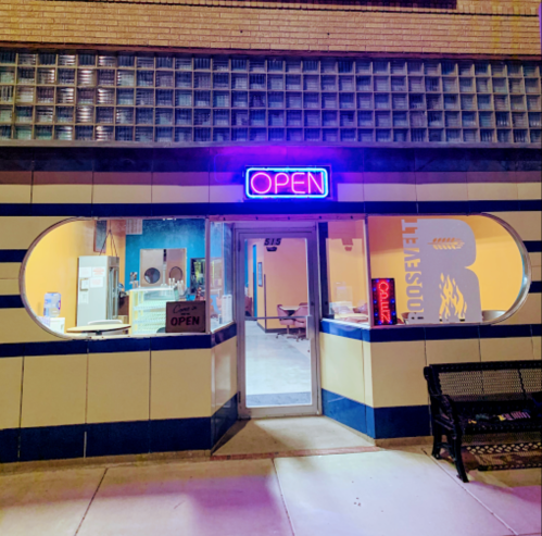 A brightly lit storefront with a neon "OPEN" sign, featuring blue and white stripes and a welcoming entrance.