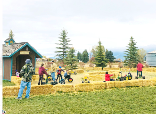Children riding tricycles on a hay bale course in a grassy area, with trees and a building in the background.