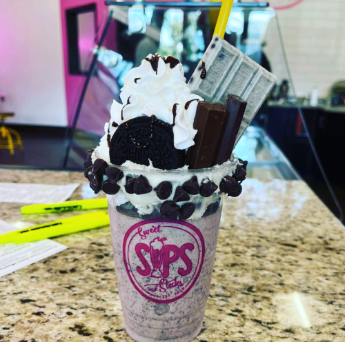 A decadent milkshake topped with whipped cream, chocolate bars, an Oreo, and chocolate chips in a branded cup.