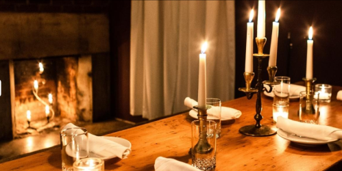 A cozy dining table set with candles and glasses, illuminated by a warm fireplace in the background.