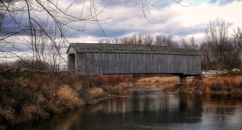 A wooden covered bridge spans a calm river, surrounded by autumn foliage and cloudy skies.