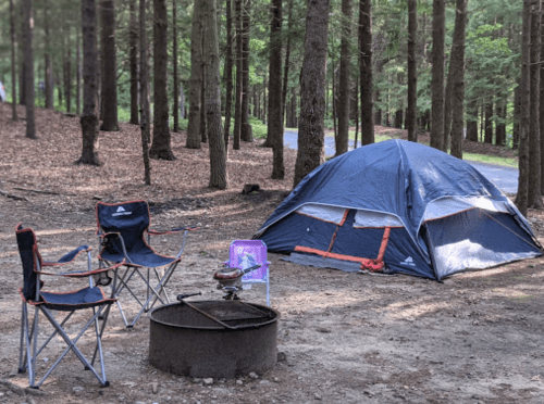 A camping scene with a tent, two chairs, and a fire pit surrounded by trees in a wooded area.
