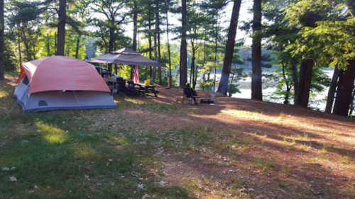 A campsite by a lake, featuring a tent, chairs, and a fire pit surrounded by trees in a sunny setting.