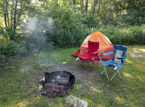 A campsite with a tent, two folding chairs, and a smoky fire pit surrounded by greenery.