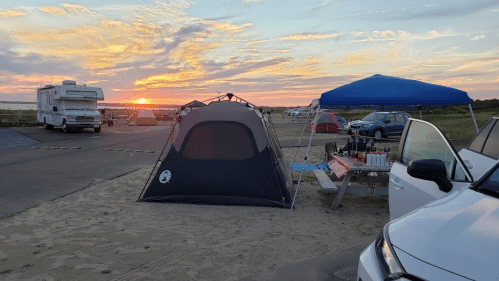 A campsite at sunset featuring a tent, a blue canopy, picnic table with food, and parked vehicles.