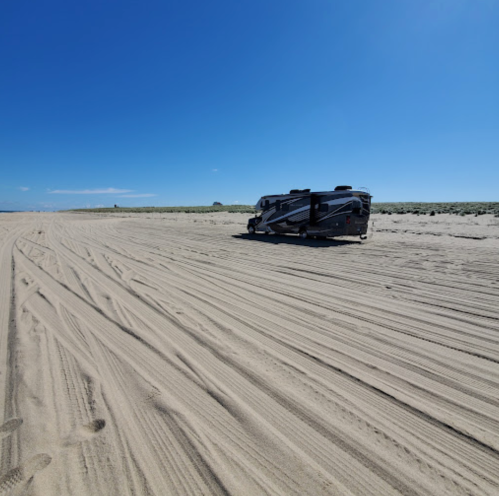 A parked RV on a sandy beach with clear blue skies and distant dunes in the background.
