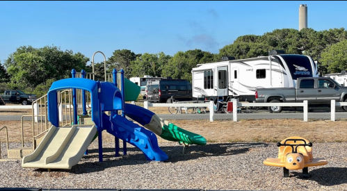 A colorful playground with slides and a small ride-on toy, set in a campground with RVs in the background.
