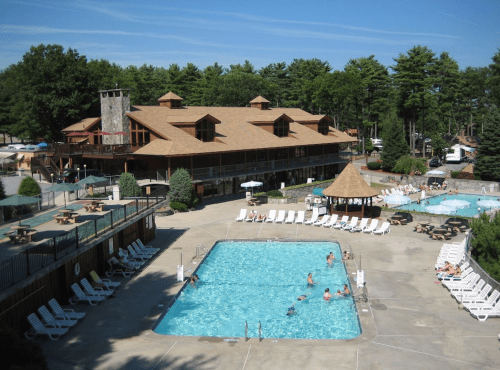 A sunny resort pool area with lounge chairs, a large pool, and a lodge surrounded by trees. People are enjoying the space.