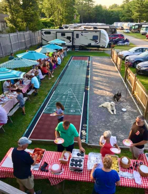A lively outdoor gathering with tables of food, a shuffleboard court, and people enjoying a sunny day.