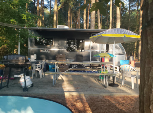 A camping setup featuring a trailer, picnic table, chairs, and an umbrella in a wooded area.