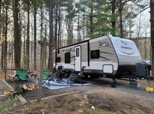 A camper parked in a wooded area, with chairs set up around a fire pit and a rug on the ground.