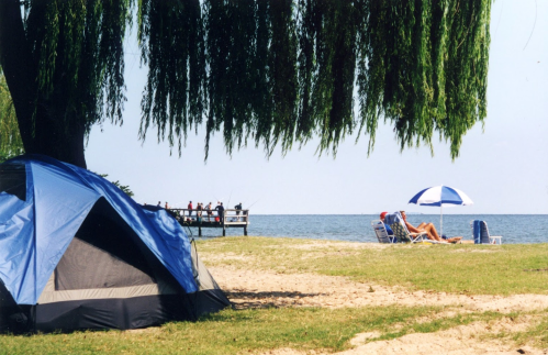 A blue tent on a sandy beach with a pier in the background and people relaxing under umbrellas.
