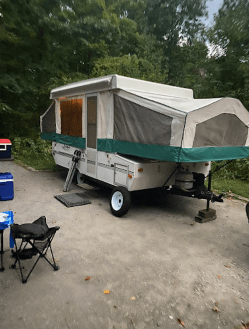 A pop-up camper parked on a gravel site surrounded by trees, with a cooler and camping chair nearby.