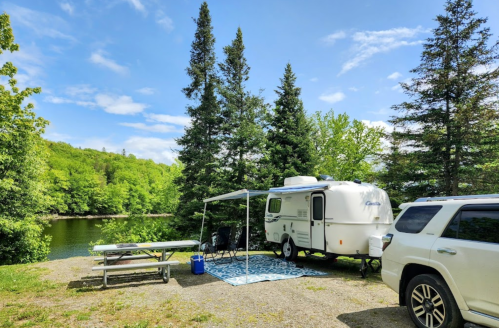 A cozy camper parked by a serene lake, surrounded by lush green trees and a picnic area.