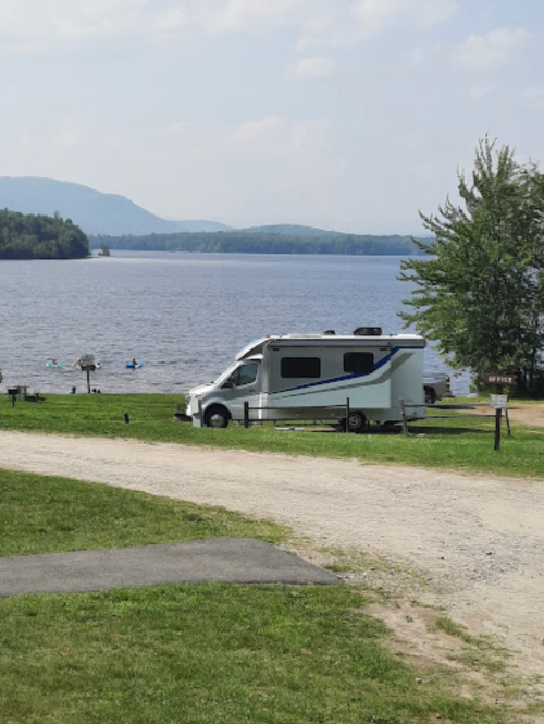 A white RV parked by a lake with mountains in the background and people kayaking in the water.
