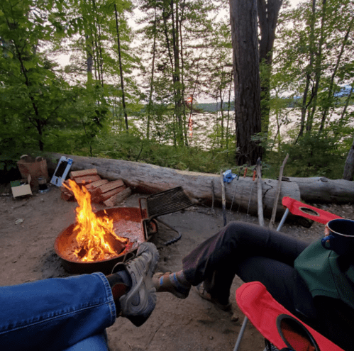 Two people sit by a campfire, with a lake and trees in the background, enjoying a cozy outdoor evening.