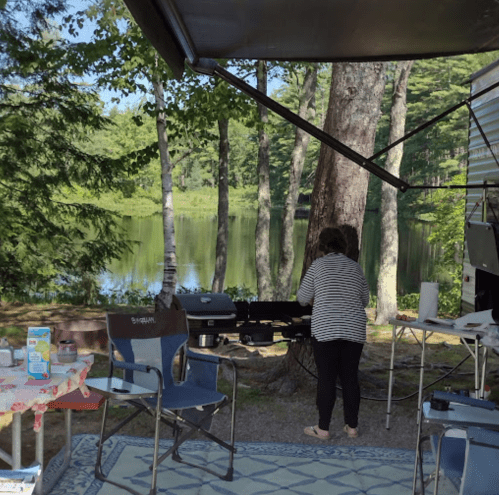 A person stands by a grill at a campsite near a serene lake, surrounded by trees and outdoor furniture.