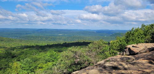 A panoramic view of lush green hills and valleys under a partly cloudy sky from a rocky overlook.