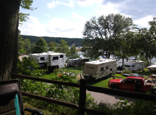 A scenic view of RVs parked by a lake, surrounded by trees and greenery, with a red truck in the foreground.