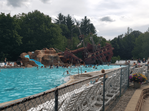 A large pirate ship play structure beside a pool, with people swimming and lounging in a lush green setting.