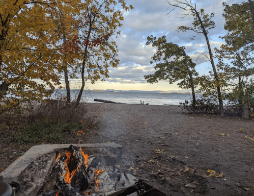 A cozy campfire on a sandy beach, surrounded by autumn trees and a serene lake under a cloudy sky.