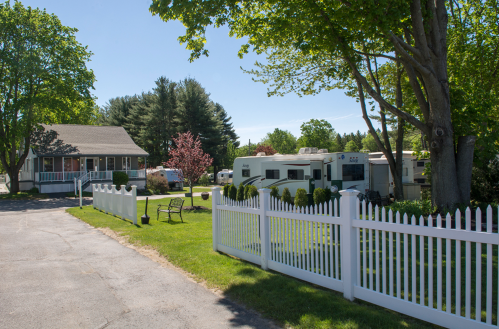 A sunny scene featuring a house and RVs beside a white picket fence, surrounded by green trees and grass.