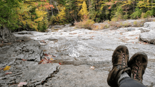 A pair of hiking boots resting on a rocky surface by a stream, surrounded by autumn foliage.