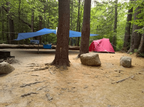 A campsite in a forest with a red tent, blue tarps, and a picnic table surrounded by trees and rocks.