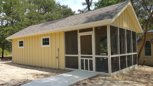 A small yellow cabin with a screened porch, surrounded by trees and a sandy area.
