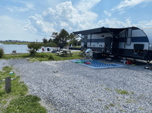 A camper parked by a lake, with picnic tables and a blue blanket on gravel, under a partly cloudy sky.