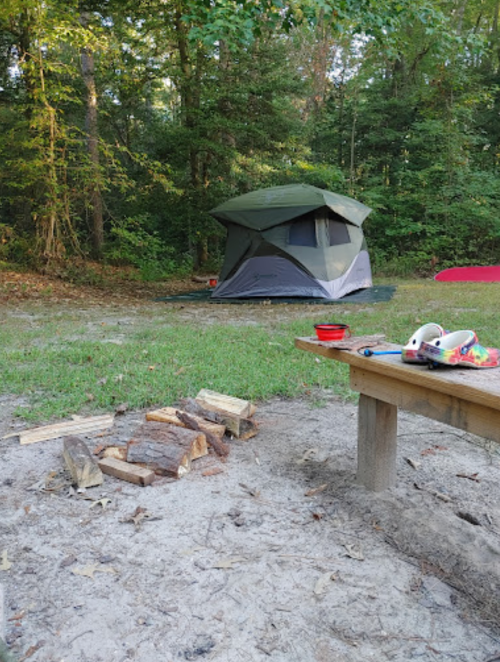 A campsite with a tent, a firewood pile, and a picnic table in a wooded area.
