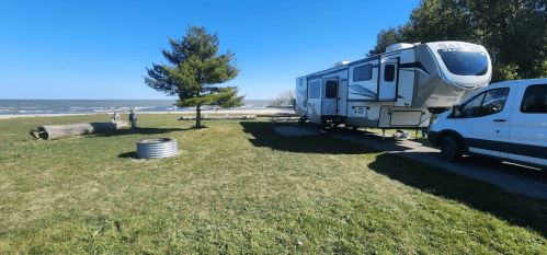A parked RV by the beach, with a van nearby and people walking along the shore under a clear blue sky.