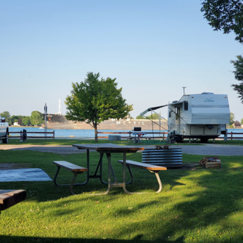 A peaceful campsite by the water with an RV, picnic table, and trees under a clear blue sky.